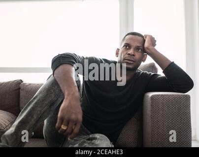 Dreaming adult black man leaning on hands while sitting on couch and looking away in shiny window Stock Photo