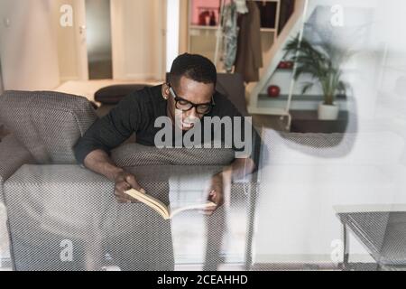 Adult African American man in glasses reading book while sitting on sofa at home Stock Photo
