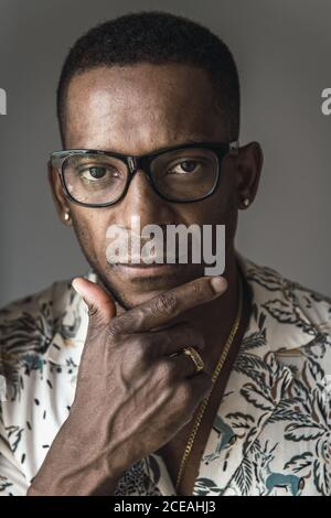 Head shot of serious adult African American man in golden accessories and glasses touching chin