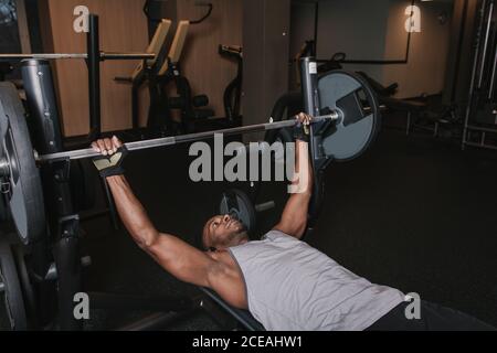 Handsome African?American man in sportswear lying on bench and lifting heavy barbell while training in modern gym Stock Photo