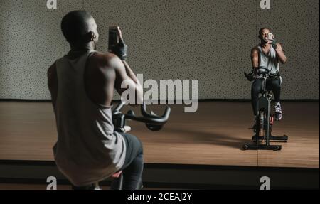 Back view of African American man taking selfie while sitting on exercise bike in front of mirror in modern gym Stock Photo