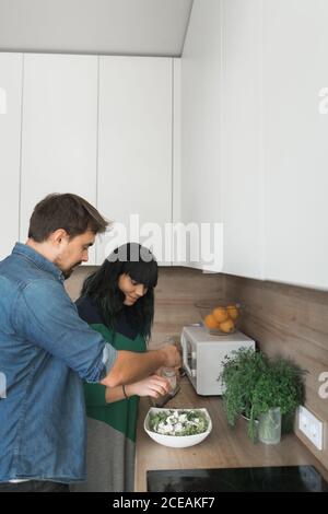 Side view of handsome young guy and lovely Woman cooking healthy salad while standing in stylish kitchen together Stock Photo