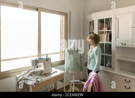 Senior female adjusting beautiful ornamental garment on dummy while working in cozy workshop Stock Photo
