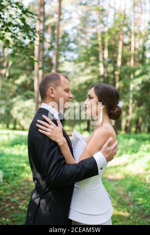 Side view of handsome groom and beautiful bride hugging and looking at each other while standing on blurred background of nature Stock Photo