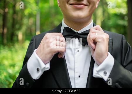 unrecognizable young man in suit smiling and adjusting bow tie while standing on blurred background of nature Stock Photo
