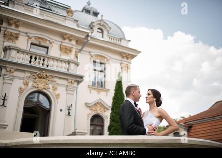 Side view of handsome groom and lovely bride looking at each other and hugging while standing near amazing luxury building Stock Photo