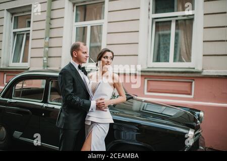 Side view of handsome young groom and pretty bride hugging while standing on city street near vintage vehicle Stock Photo
