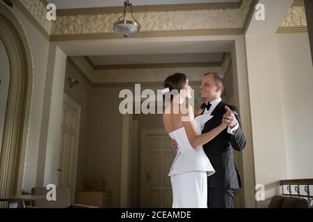 Side view of attractive bride and groom smiling and looking at each other while dancing inside beautiful luxury building Stock Photo
