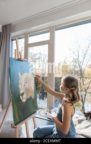 Side view of little talented girl drawing painting sitting at easel against window at home Stock Photo