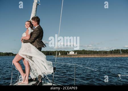 Side view of happy married couple standing on yacht nose and embracing on lake Stock Photo