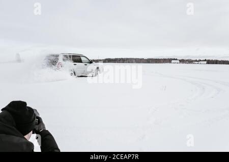 Back view of crop guy shooting on camera automobile drifting on snow meadow and cloudy sky in Vilnius, Lithuania Stock Photo