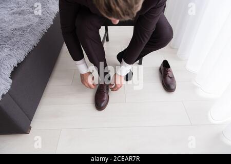 Young man in black costume and white shirt sitting on chair in bedroom and putting on leather shoes Stock Photo