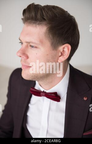 Young handsome man in black formal costume and white shirt with stylish hairdo sitting in room and looking at camera with hand on chin Stock Photo