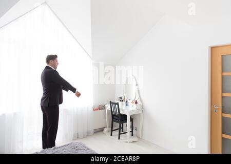 Side view of young handsome man standing in white bedroom and putting on black formal costume Stock Photo