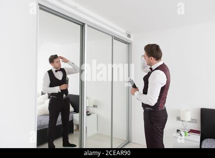 Side view of young handsome man in trousers and waistcoat standing in bedroom and looking in mirror Stock Photo