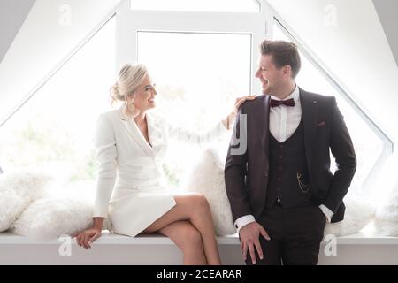 Young attractive Woman in white jacket sitting on window sill with handsome man in black costume standing near and smiling Stock Photo