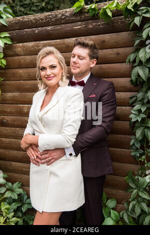 Elegant adult bride and groom embracing while standing in suits against wooden wall and smiling at camera Stock Photo