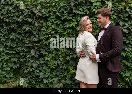 Elegant adult bride and groom embracing while standing in suits against wooden wall and smiling at camera Stock Photo