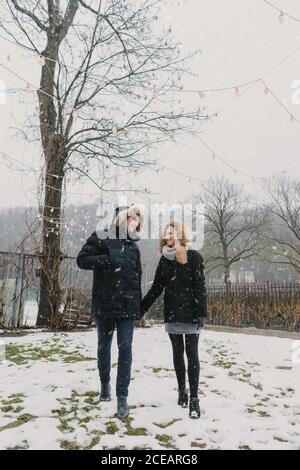 Side view of happy guy and young lady in ski jackets holding hands and walking on the snowy street Stock Photo