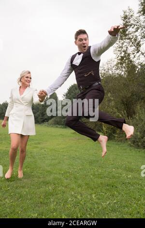 Trendy adult groom and bride in elegant suits holding hands and jumping with excitement on green meadow Stock Photo