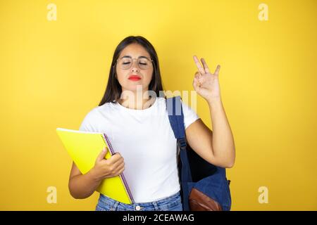 Young beautiful student woman wearing backpack holding notebook over isolated yellow background relax and smiling with eyes closed doing meditation ge Stock Photo