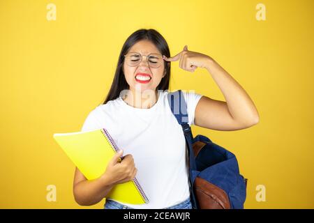 Young beautiful student woman wearing backpack holding notebook over isolated yellow background with hand on head for pain in head because stress. Suf Stock Photo