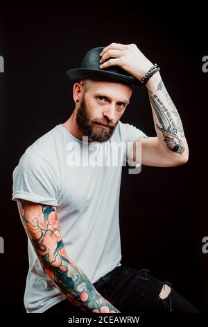 Cheerful bearded hipster in hat and t-shirt with tattoos on arms on black background Stock Photo