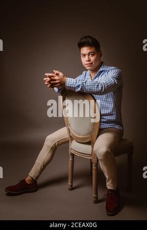 Young man in shirt sitting on chair and looking at camera on dark background Stock Photo