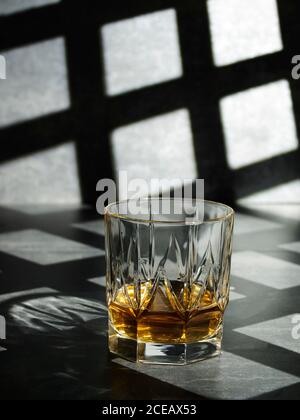 Close-up of clear crystal glass filled with golden liquor on gray background in shadow of grid Stock Photo