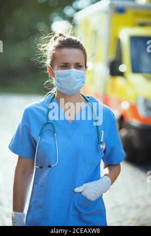 coronavirus pandemic. modern paramedic woman in scrubs with stethoscope and medical mask outdoors near ambulance. Stock Photo