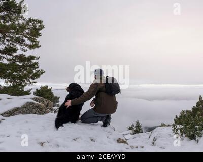 Hiker and his dog looking in the snowy mountains at foggy sky background. Stock Photo