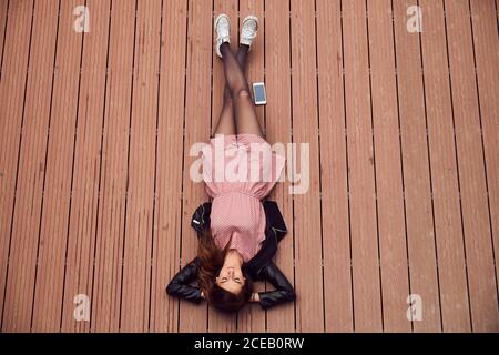 From above view of young attractive Woman in leather jacket and dress lying on striped surface with mobile phone placed near Stock Photo