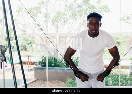 Muscular African American sportsman doing cable fly with exercise machine standing against window during training in gym. Stock Photo