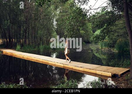 Back view of young Woman in dress with long hair walking on wooden bridge above calm smooth river in forest Stock Photo