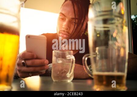 Crop view of young African-American guy holding and looking at mobile phone, sitting in cafe near glasses with beer in Austria Stock Photo