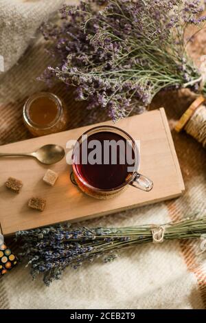 Bunches of dried herbs and jar of fresh honey lying on blanket near board with cup of hot tea and cubes of sugar Stock Photo