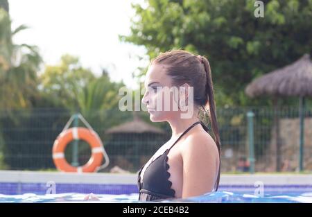 Side view of young Woman in black swimsuit standing in pool and looking away Stock Photo