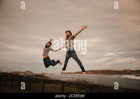Young happy Woman and man in hats holding hands and having fun on seat on coast near waving sea and cloudy sky Stock Photo