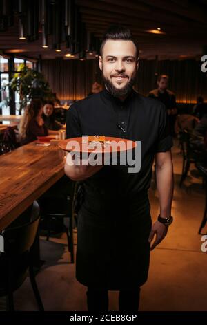 Cheerful bearded waiter with a slice of cake in a Georgian restaurant. Beautiful waiter in black clothes with a beard and a serving of cake Stock Photo