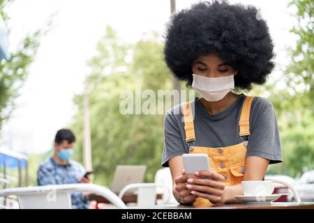 African lady wearing face mask using phone sit at table in outdoor cafe. Stock Photo