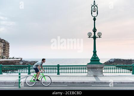 Side view of young sporty man riding bicycle on bridge at the oceanside Stock Photo