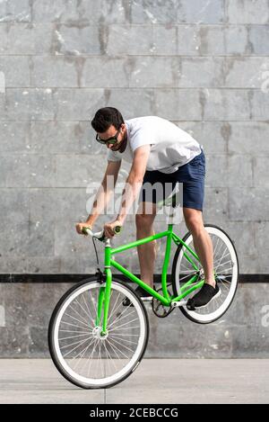 Young man in sunglasses performing trick and balancing on front wheel of green bicycle Stock Photo