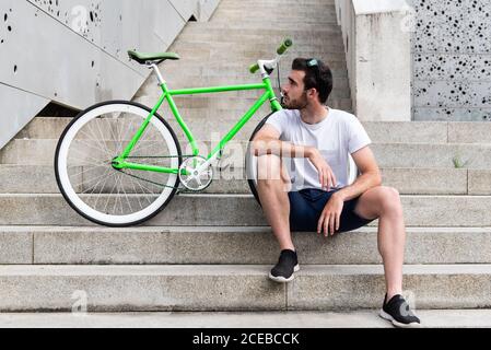 Young man sitting on steps at parked green bicycle on street Stock Photo