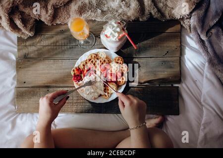 Young Woman eating appetizing waffles and tasty drinks on tray on bed Stock Photo
