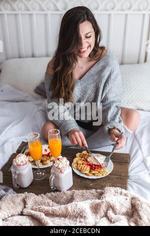 Young Woman eating appetizing waffles and tasty drinks on tray on bed Stock Photo