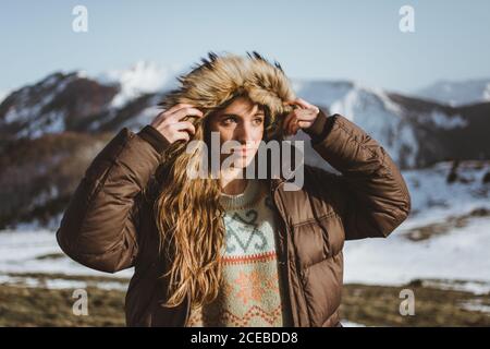 Attractive Woman putting on hood of warm jacket and looking away in snowy hills. Stock Photo