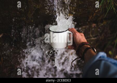 Closeup shot of hand of anonymous man holding mug under fresh water of cold water source in nature Stock Photo