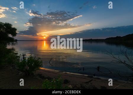 Sun and rays of sunlight peeking out from below clouds at sunrise on a calm lake with trees and a bit of sandy beach. Stock Photo