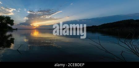 Panoramic view of sunlight and clouds reflecting on the smooth surface lake with trees lining the shores on both sides at sunrise. Stock Photo