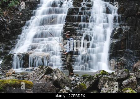 Fisherman dressed in fishing hat with blue shirt and gray marching trousers holding a spinning rod standing on rocks behind the slope of a waterfall Stock Photo